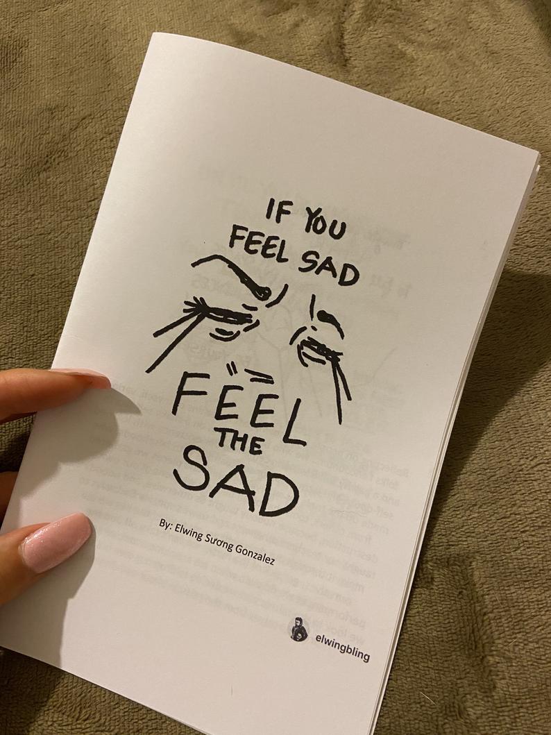 photo of zine: If You Feel Sad, Feel the Sad, held by a mostly off-camera hand. Cover has a handwritten title and drawing of crying eyes/shoulder & torso