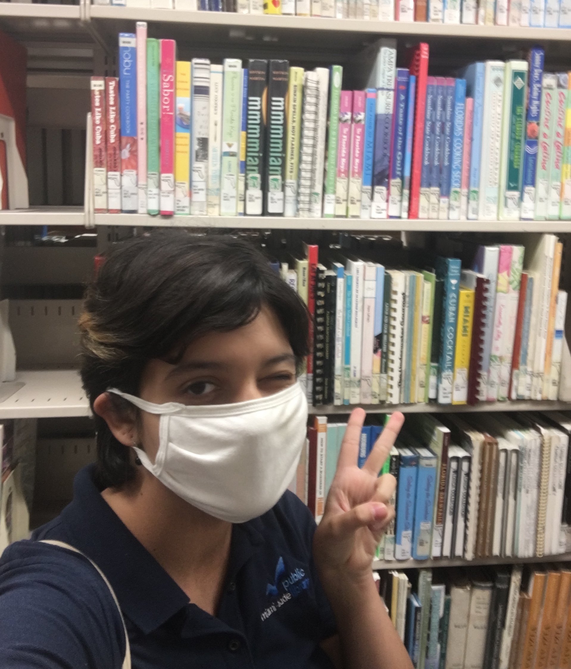 photo of person in a white mask in front of a bookshelf holding hand in a peace sign and winking