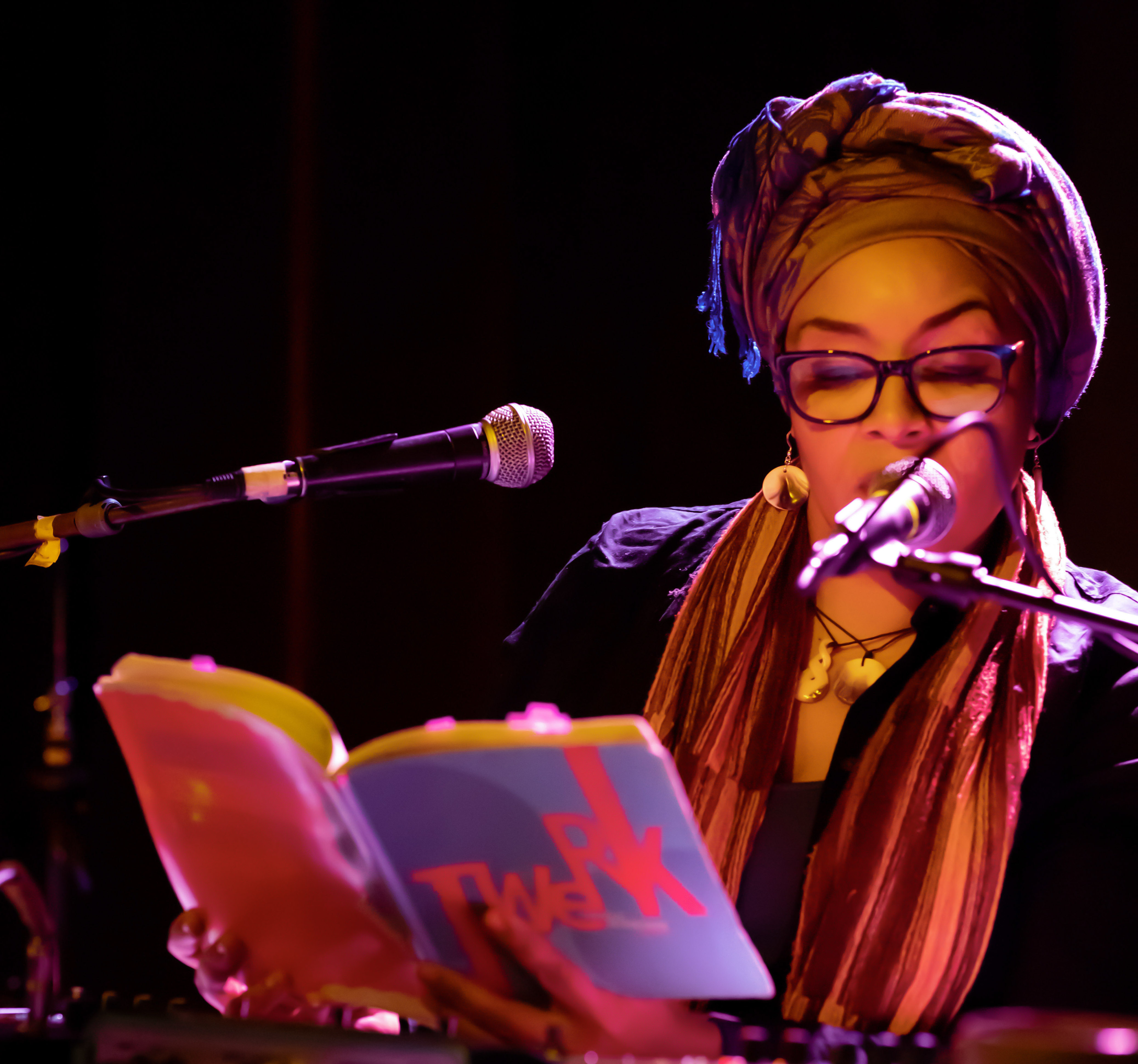 A woman reads from a book on a stage with two microphones.