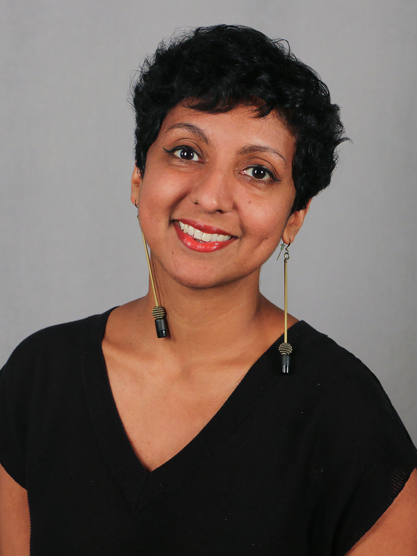 A South Asian person with short black hair wearing a black V-neck smiles into the camera.