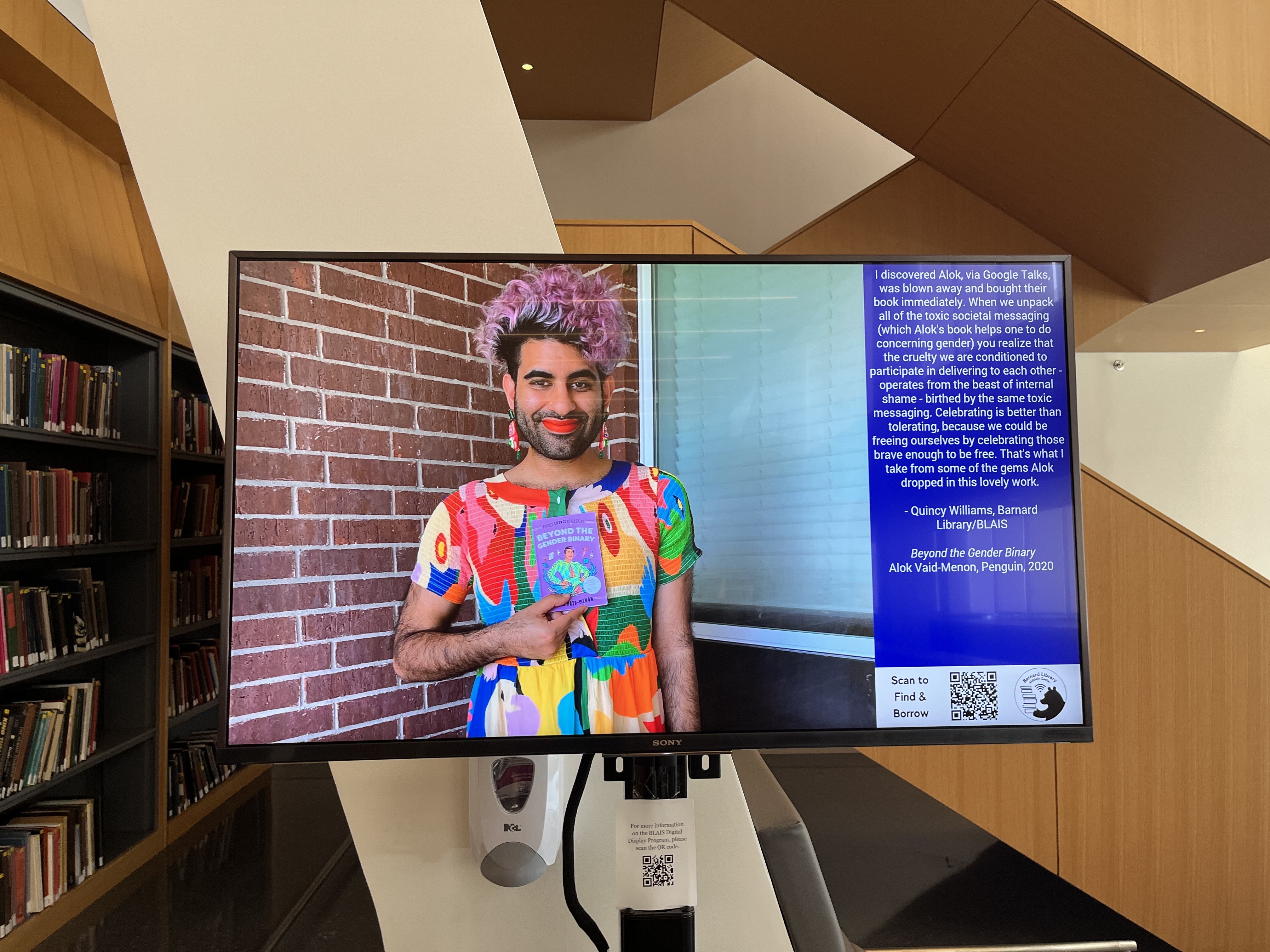 Digital screen in Milstein Lobby, displaying a photo of Alok Viad-Menon holding their book Beyond the Gender Binary. Alok is wearing a brightly patterned dress, as well as brightly colored earrings. They have a beard, pink hair, and are wearing red lipstick. To the right of the photo, there is text about the book from Quincy Williams.