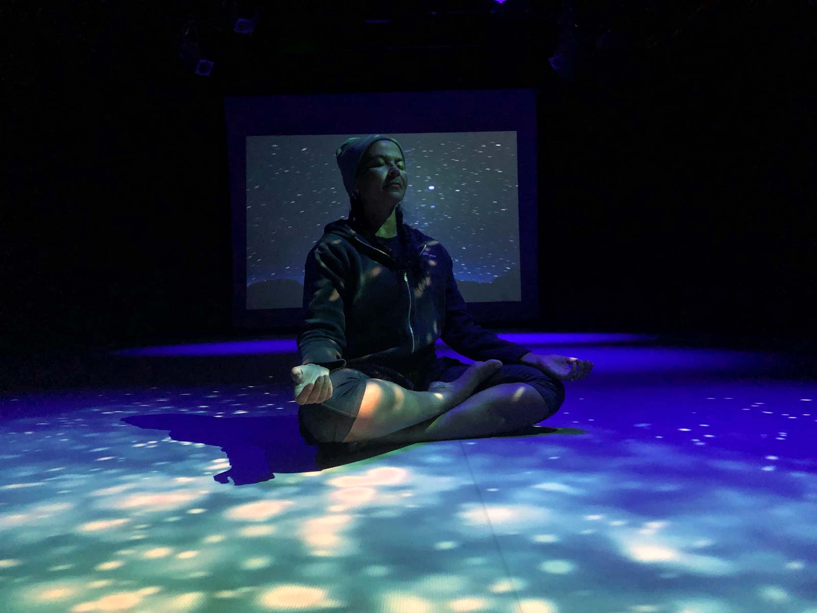 woman sits cross-legged on the floor, eyes closed, in a dark room