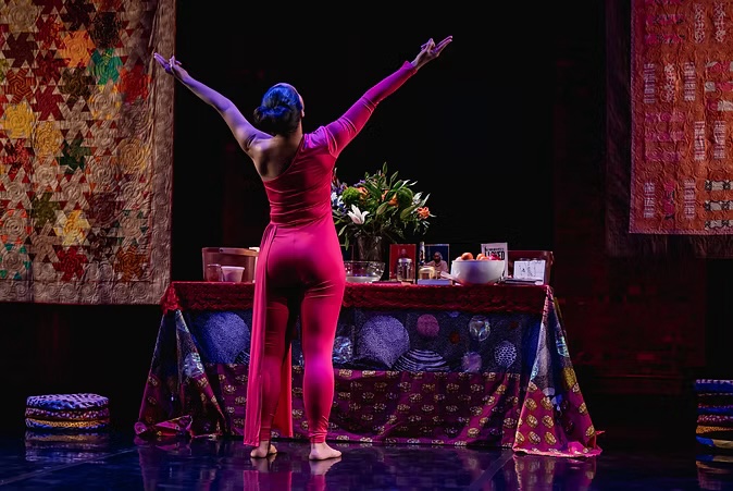 Image from Purple depicts a dancer with arms outstretched in front of an altar and hanging quilts. Photo by Effy Grey