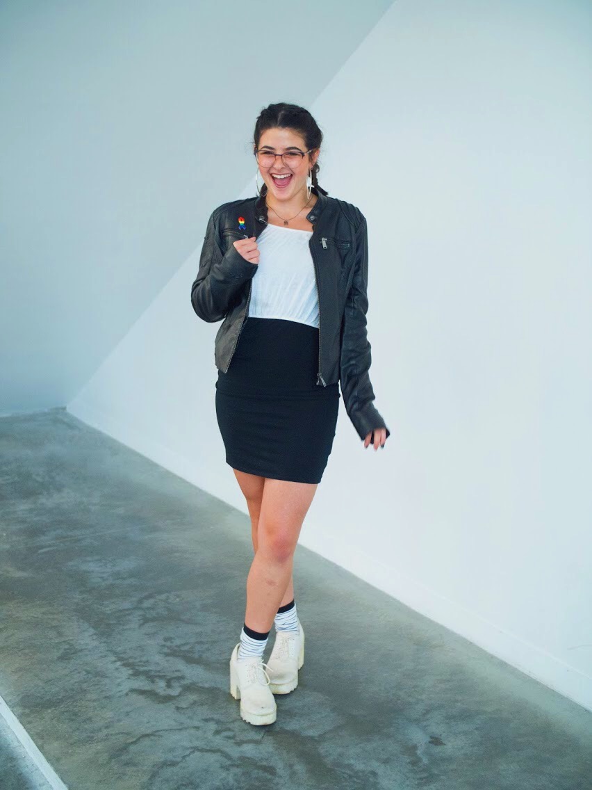photo of Alisia in a leather jacket, white shirt, black mini-skirt, and white platforms. She is wearing braids and big silver hoops and has a big smile on her face.