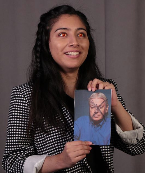 A student with glowing red eyes grins and holds up a portrait of a man with his face X'ed out.