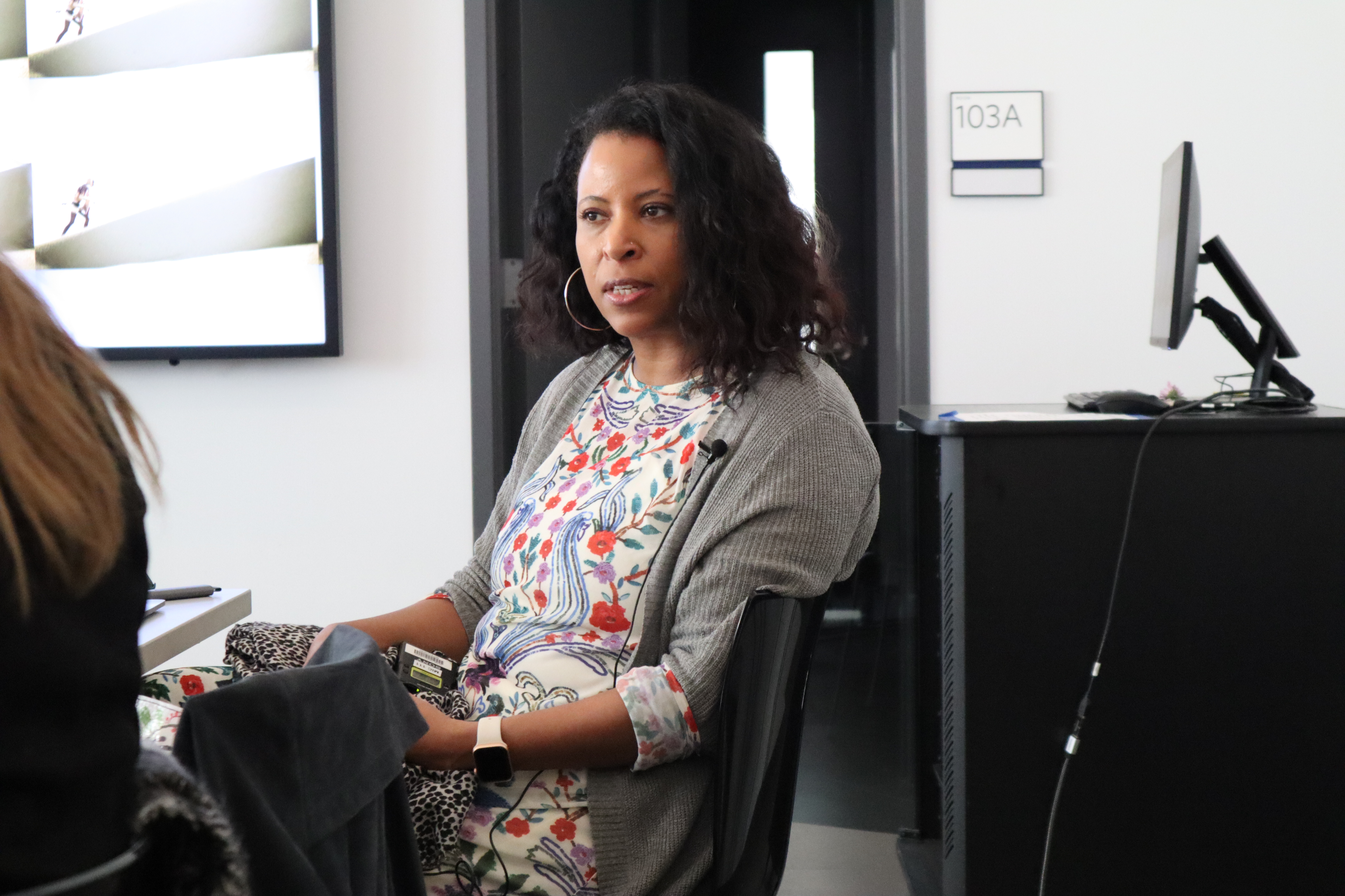 Marisa Parham, wearing a white dress with colorful embroidery all over it and a grey cardigan, mid-talk.