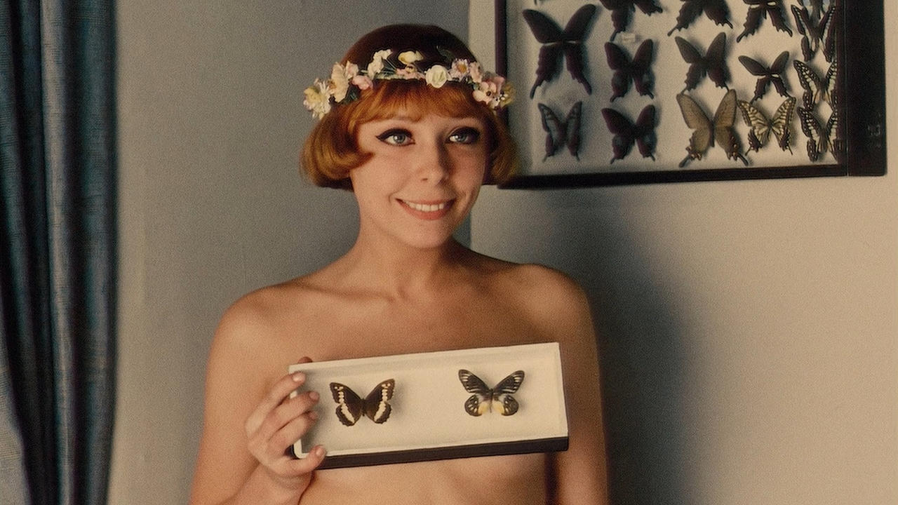 A woman with a flower crown smiling mischievously. She holds a picture frame with two butterflies in it over her chest.