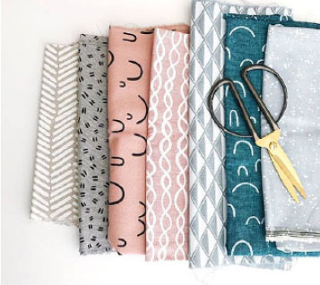 A pile of assorted patterned fabrics in light pink, teal, and gray, with a pair of fabric scissors on top. 