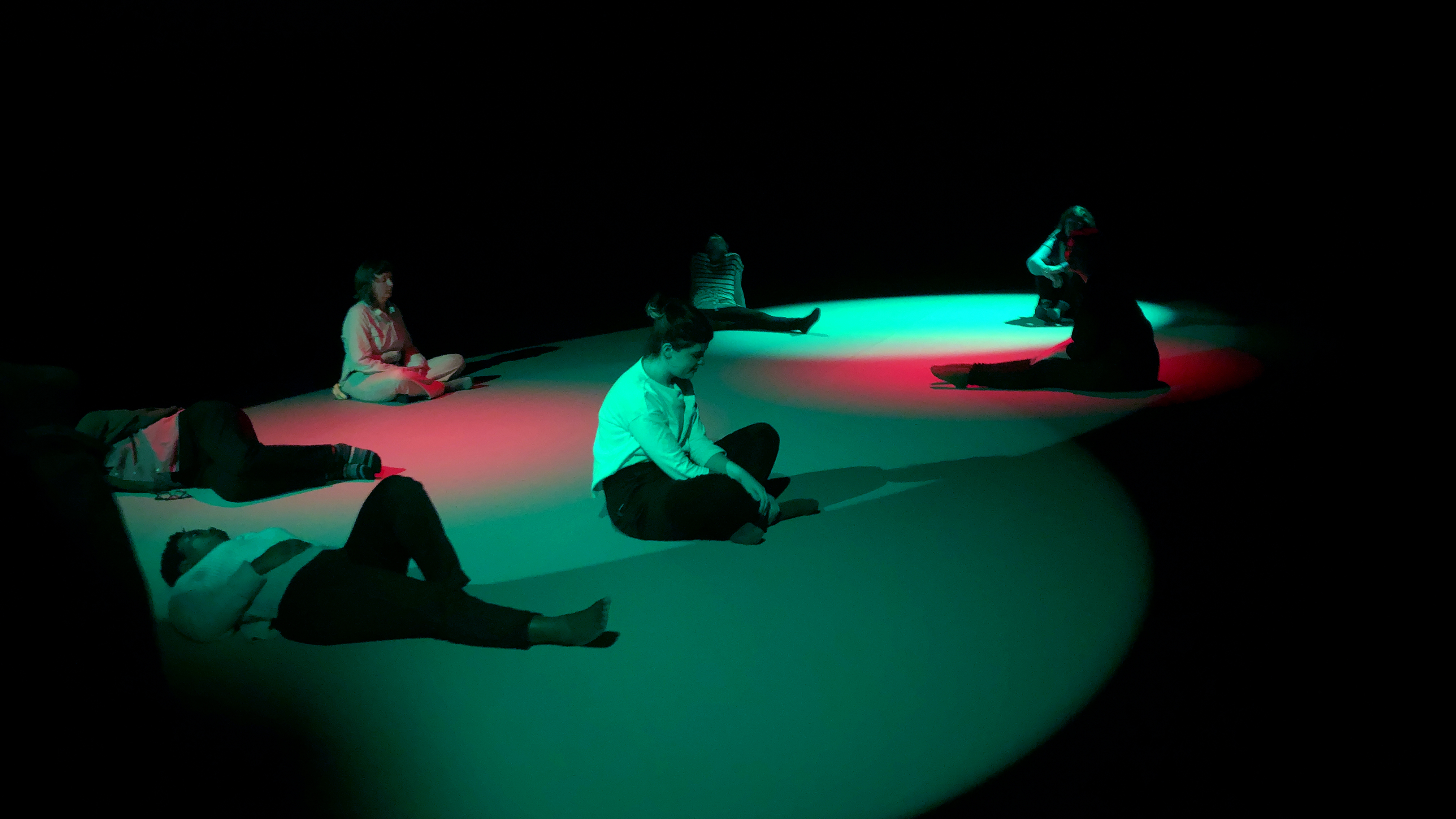 Students and participants sit contemplatively on the floor, which is covered in cyan and red light.