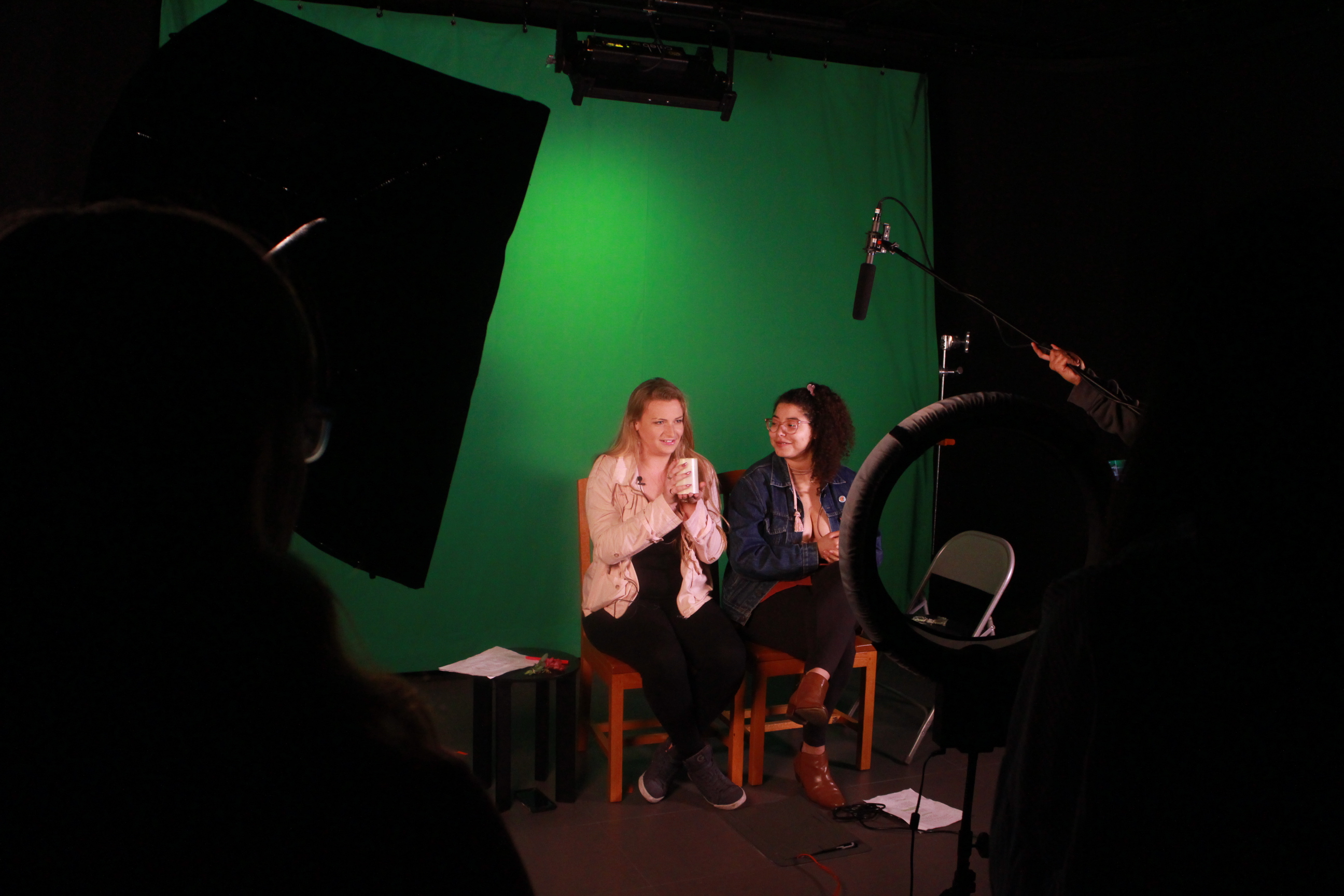 Two students, acting as beauty vloggers, sit on wooden chairs in front of a green screen