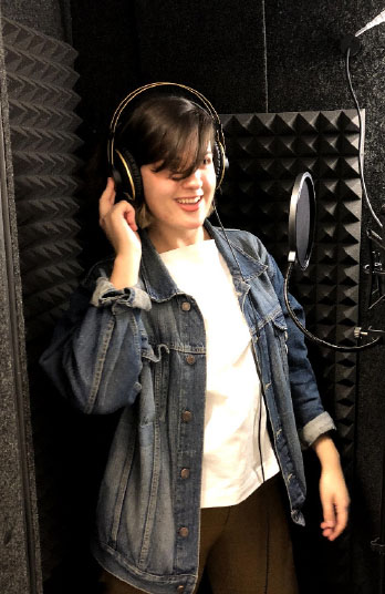 Ruby, wearing black headphones, speaks into a microphone inside the padded audio recording booth.