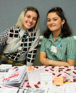 Two students are tabling at 2018's Feminist Zine Fest, with their zines arranged on their table. 