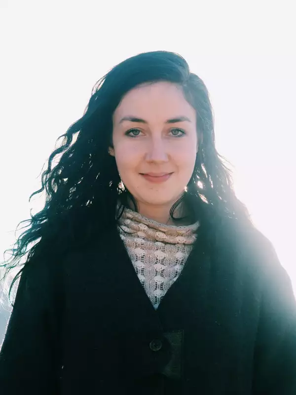 Woman outdoors with long brown curly hair and green eyes, wearing a sweater and jacket