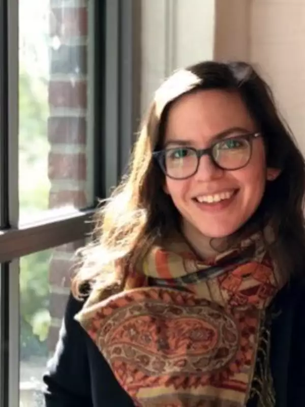 Woman with long brown hair, wearing glasses and a patterned scarf, standing by a window